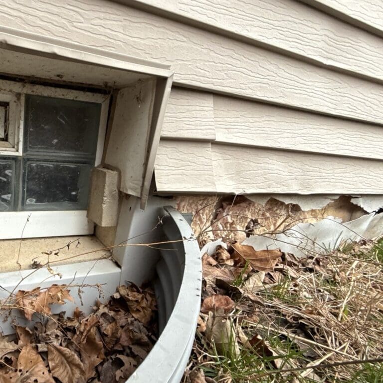 Close-up of deteriorated exterior siding near a window showing cracks, warping, and exposed underlying material with debris buildup