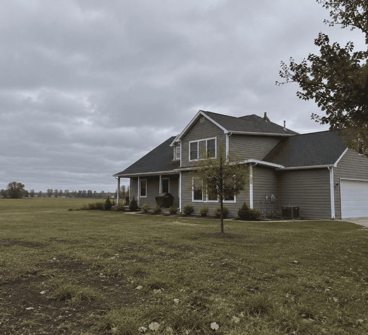 Rural Wisconsin home with neutral siding surrounded by open fields and exposed landscape under an overcast sky.